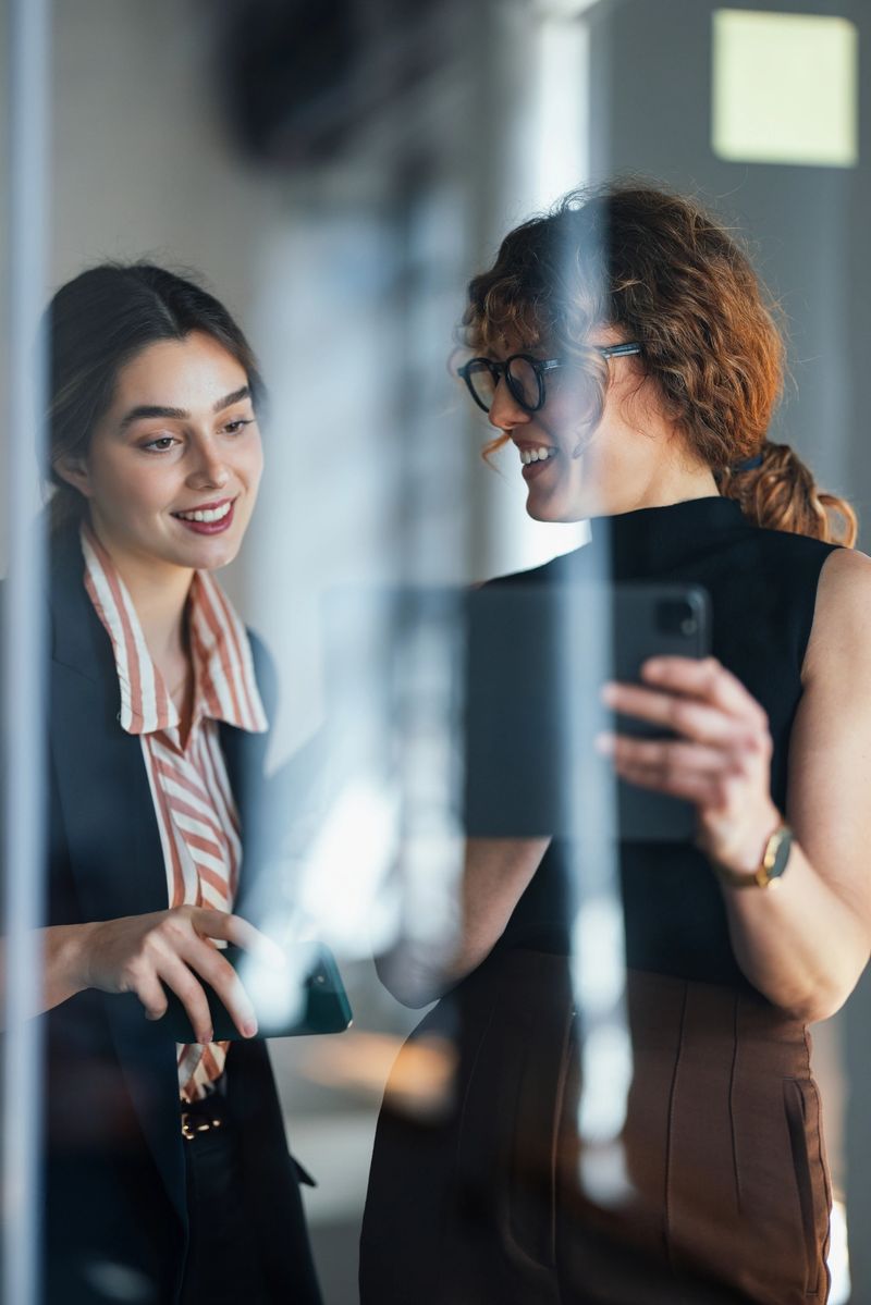 Two professional women discuss a project in a bright office, sharing a smile while looking at a smartphone. The scene conveys collaboration, networking, and modern teamwork in a coworking environment.