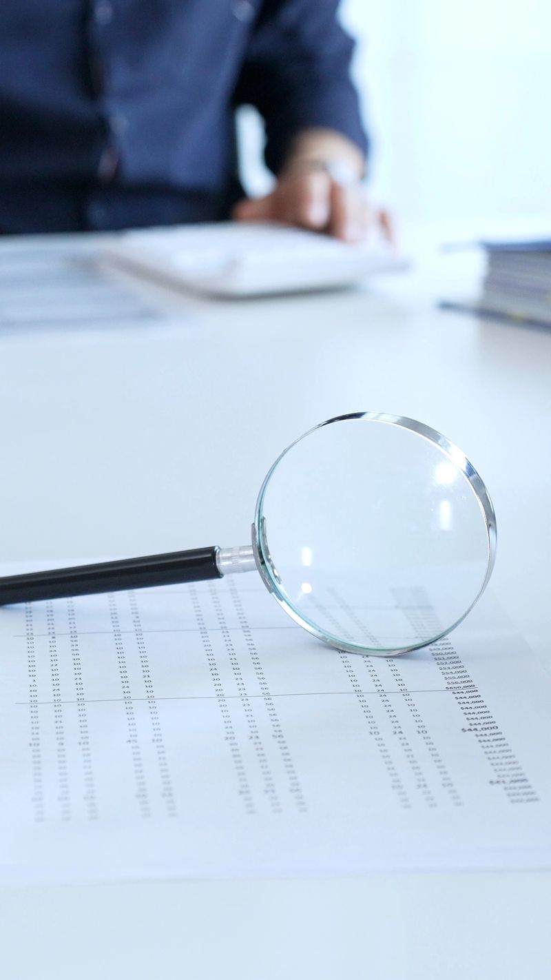 Magnifying glass resting on a financial report, with a businessman focused on a laptop in the blurred background, emphasizing financial analysis and due diligence. Audit and taxes theme in business