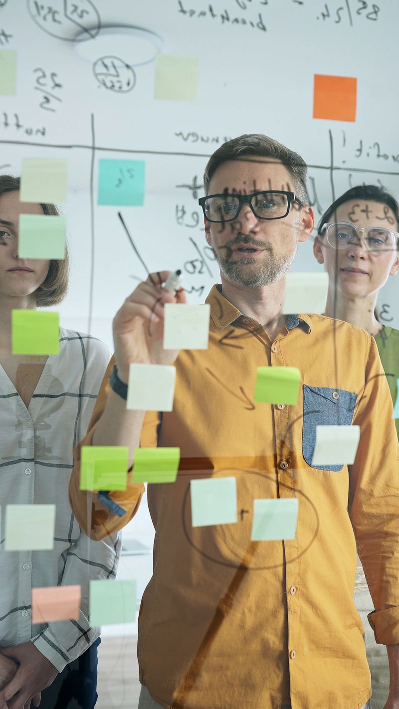 Business team collaborating on a project, using a transparent board with sticky notes and writing down ideas and plans during a productive meeting