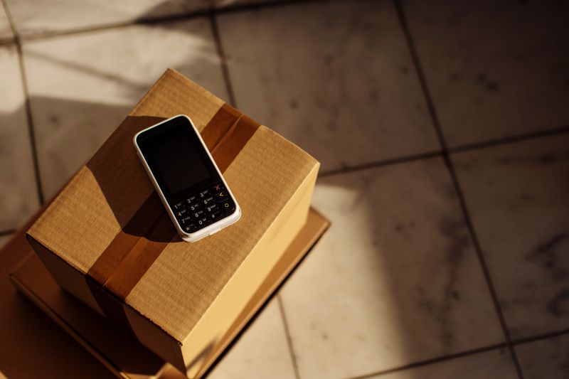 A small smartphone rests on a cardboard package bathed in warm sunlight, suggesting e-commerce, delivery, and mobile communication; Ideal for stock imagery about shipping, tech, and everyday logistics