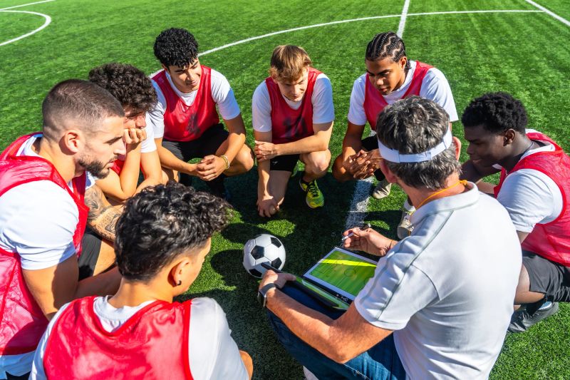 Soccer coach reviews tactics on a tablet while a diverse group of young adult players kneel in a focused huddle on a sunny green field during outdoor practice