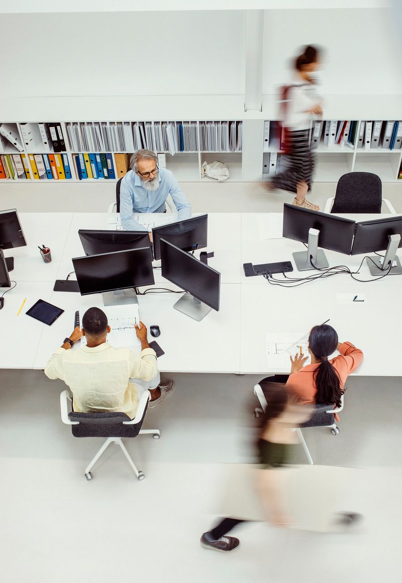 High angle view of a busy architecture biro where diverse employees are actively working at multiple computer stations. The space is organized with rows of desks, computers, and shelving filled with files, reflecting a dynamic and collaborative work environment.