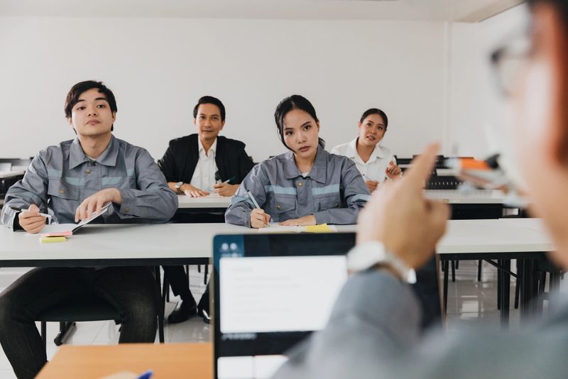 Five students are seated in a classroom, focused on an instructor who is presenting from a laptop. They appear to be taking notes and participating actively in the learning process.