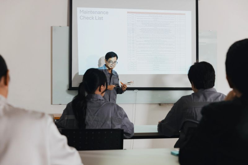 A maintenance training session is taking place in a classroom. An instructor stands at the front, presenting a checklist on a screen while students attentively listen and take notes.