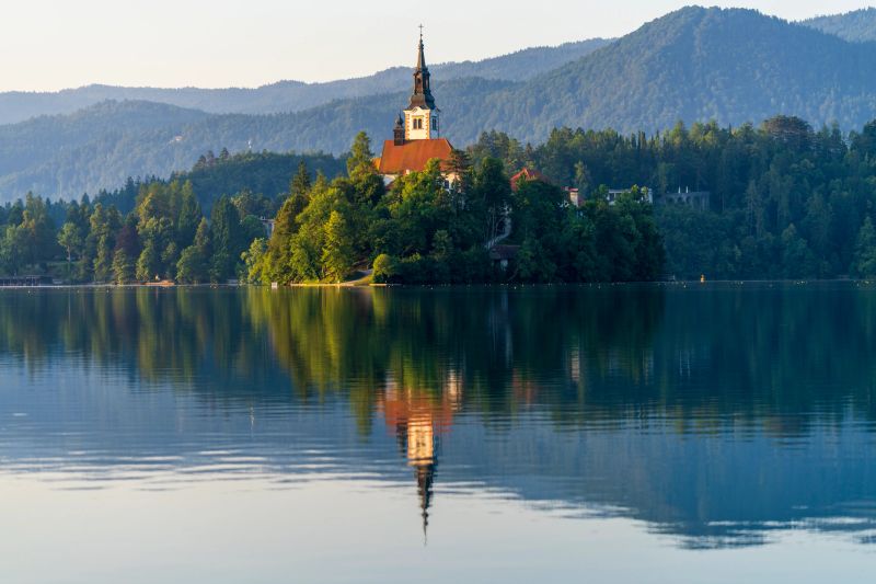 Church nestled on Lake Bled Island reflecting in tranquil water. Lush greenery surrounds the church, with mountains providing a majestic backdrop under clear skies.