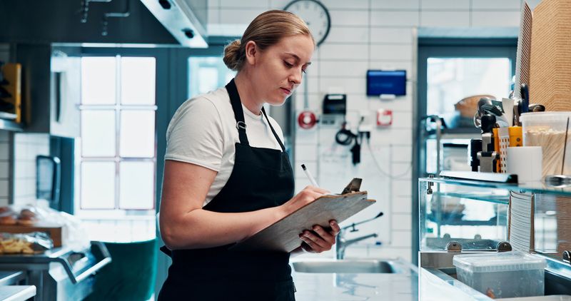 Chef, food inspection or writing in kitchen with checklist for safety, hygiene or inventory management. Woman, notes and clipboard in restaurant for quality control, hospitality and menu development.