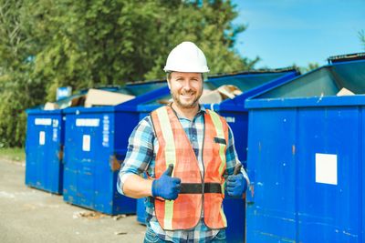 Man in safety gear giving thumbs up near blue industrial bins.
