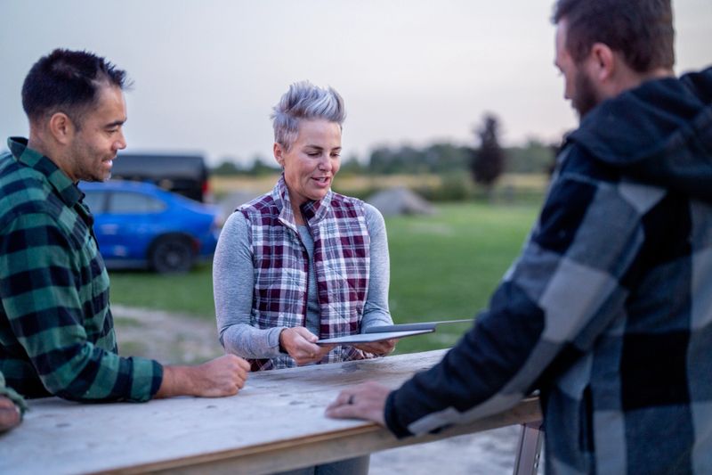 A diverse group of friends and colleagues gathers outdoors around a wooden table. A gray-haired woman hands a tablet to two men, suggesting collaboration and friendly teamwork in a relaxed setting.
