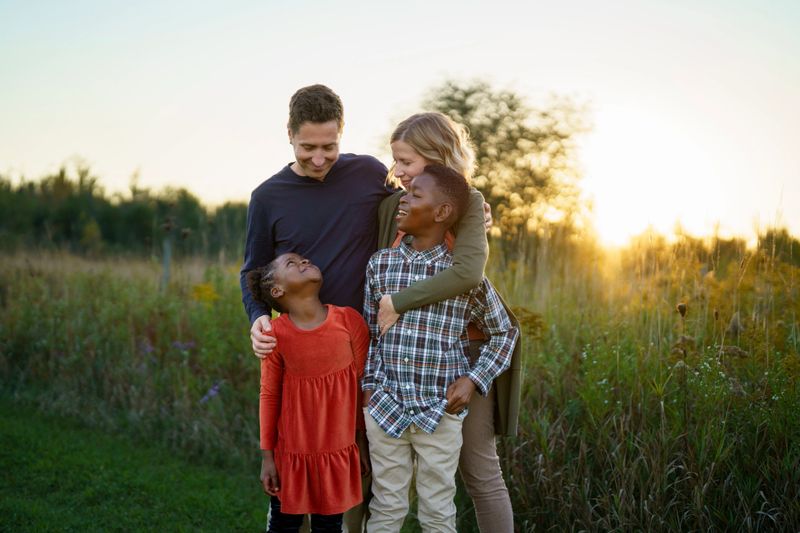 A loving family of two parents with their two children share a playful moment in a sunlit field at golden hour. The scene captures connection, warmth, and togetherness.