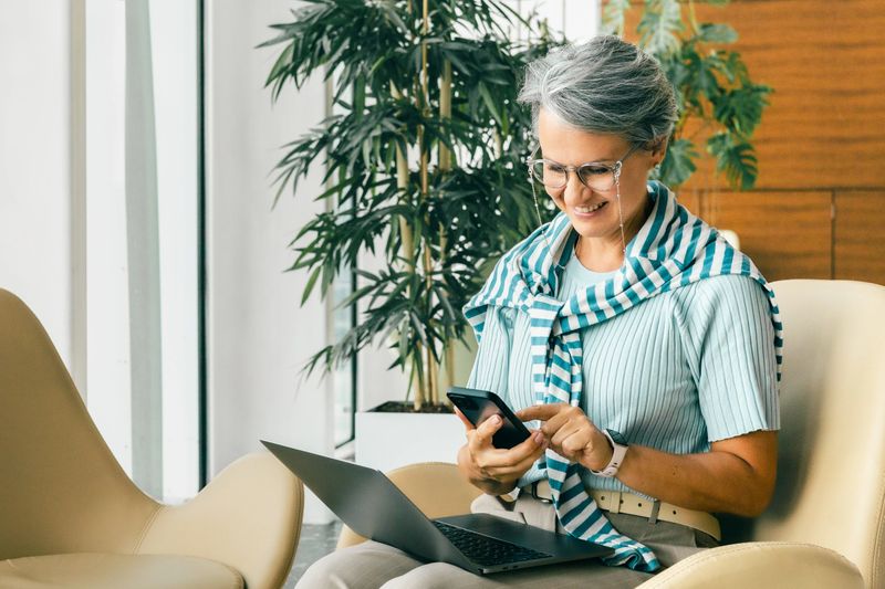 Happy mature woman multitasks with a smartphone and laptop in a bright modern space, showing productivity, confidence and a positive approach to digital communication and remote work.
