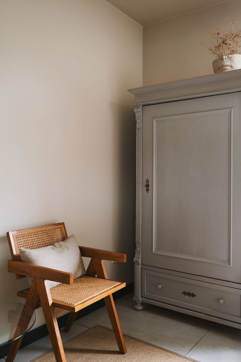 Vertical shot of inviting living room features a stylish decorative cushion on wooden chair and a gray wardrobe set against a warm toned wall, creating a serene and comfortable atmosphere in the home.