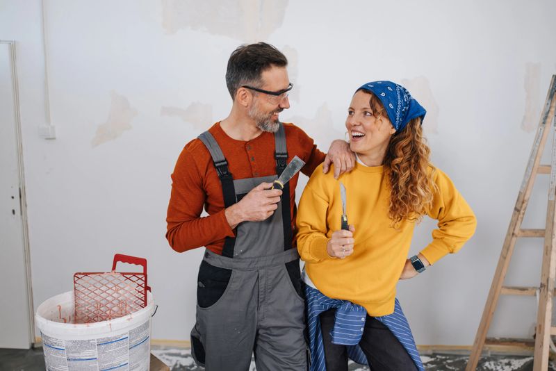 A joyful couple stands together in a room ready for renovation, sharing smiles and holding tools, reflecting teamwork and creative collaboration in home improvement.