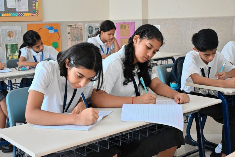 Group of Indian middle-school students sitting at their desks and focused on writing in their notebooks during class. Two girls in the front row are concentrating on their work, with other classmates studying in the background, showing a disciplined and engaged learning environment at an Indian school.