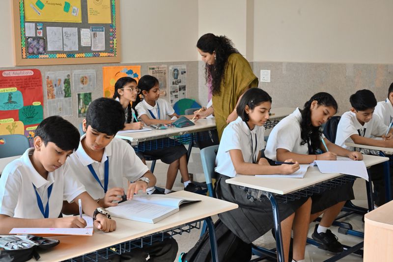 A group of Indian middle-school students sitting at their desks and working in notebooks while a female teacher in a saree walks around the classroom, assisting a girl with her lesson.
