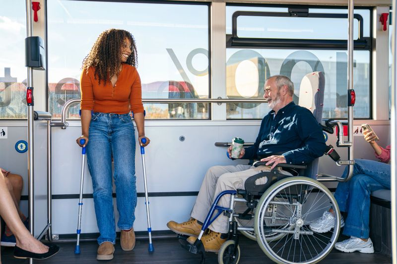 Young woman with crutches and mature adult man in wheelchair smiling and talking while traveling on a public bus