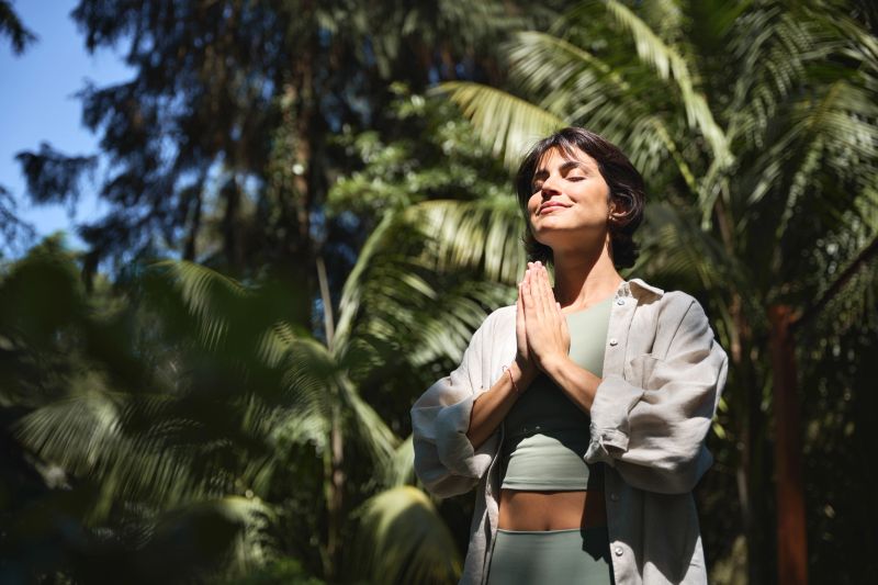 Happy calm young Hispanic woman holding hands in namaste meditating doing yoga breathing exercises with eyes closed feeling peace of mind, mental balance standing in green nature tropical park.
