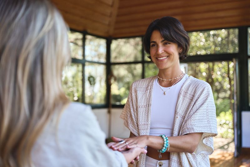 Happy female holistic teacher holding hands of woman during spiritual practice meditation experience feeling energy, peace and love, giving support during body health retreat healing training session.