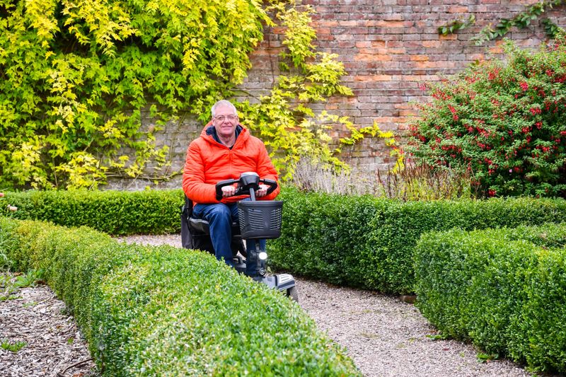 Happy man riding his mobility scooter in public park garden on an Autumn day, enjoying the freedom that the disability vehicle gives him to get out and have fun on his own, without relying on carers .