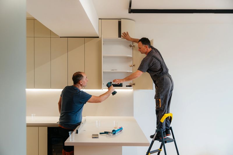Two male workers collaborate while installing upper kitchen cabinets in a modern kitchen. One man stands on a step ladder holding the cabinet door, while the other hands him a cordless drill across the kitchen counter. The bright interior, clean surfaces, and tools on the countertop highlight a professional kitchen installation process.