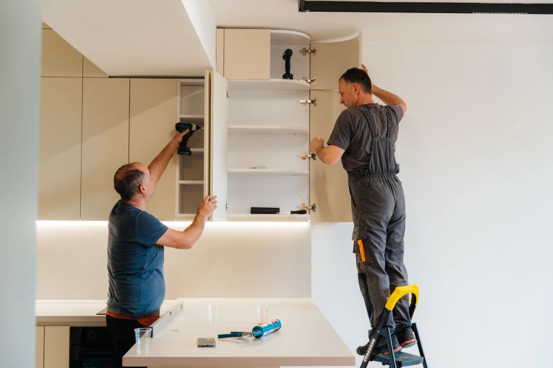 Two men are working together on the installation of upper kitchen cabinets. One man stands on a ladder adjusting the cabinet door while the other uses a cordless drill to secure the hinges. Modern kitchen interior with beige cabinets and LED strip lighting.