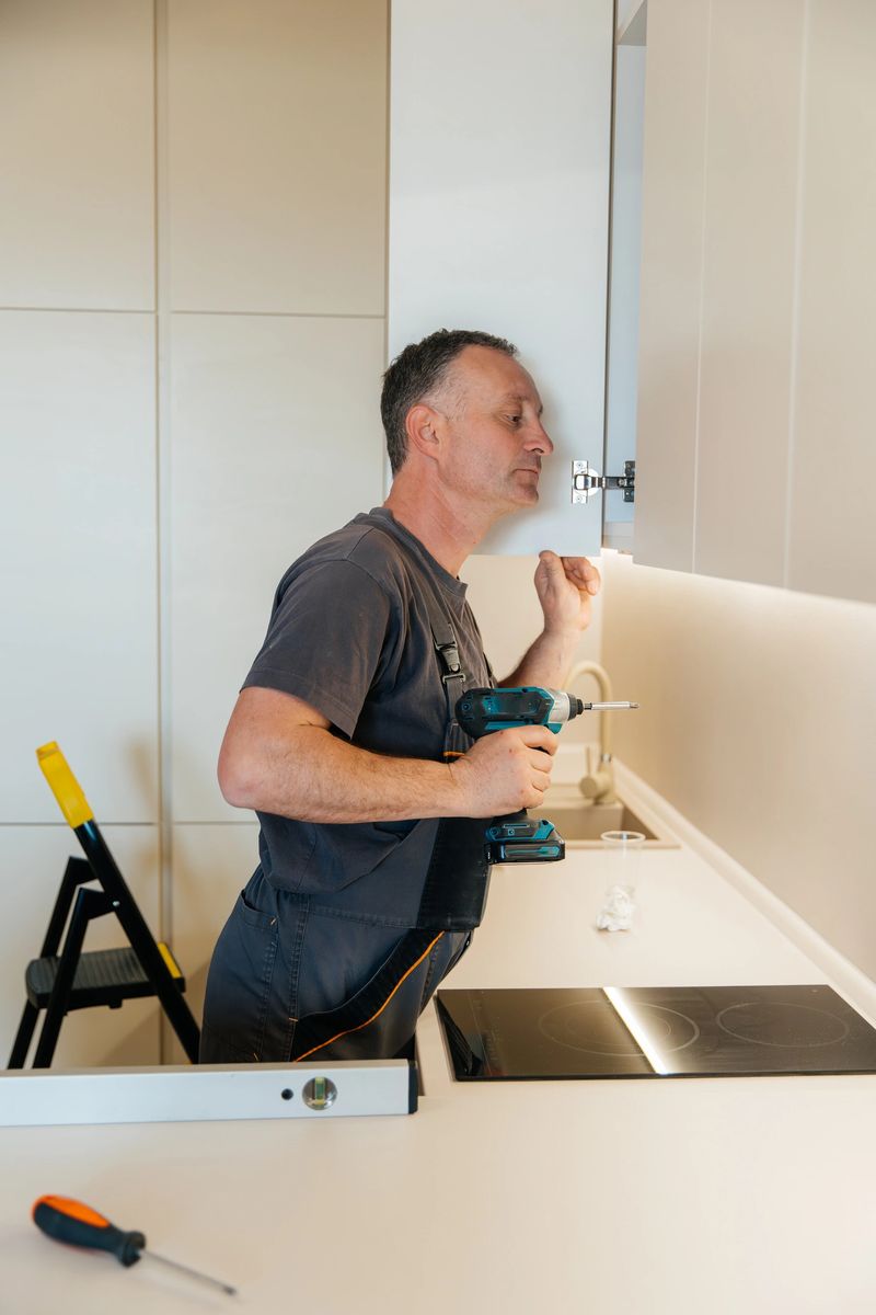 Adult handyman in grey overalls using an electric screwdriver to adjust the hinge of a modern kitchen cabinet. Concept of carpentry, repair, and home improvement indoors.