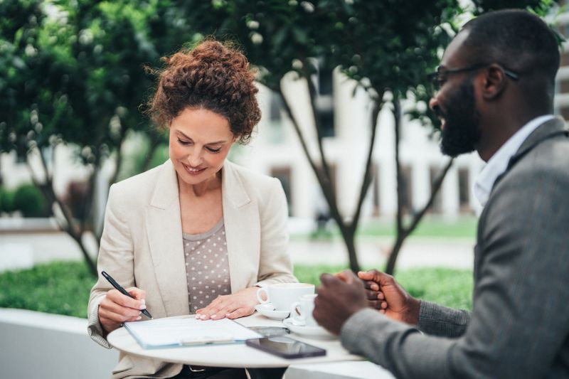 Business colleagues having serious and engaged conversation on meeting in sidewalk cafe. Real estate agent meet client in cafe, ready to sign contract. Shot of two colleagues having a discussion outdoors and preparing documents for new agreement. Confident business people working together and form filling during coffee break.