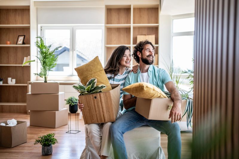 Young adult couple is smiling and looking away, holding boxes with plants and pillows, bringing belongings into their new home with empty shelves in the background