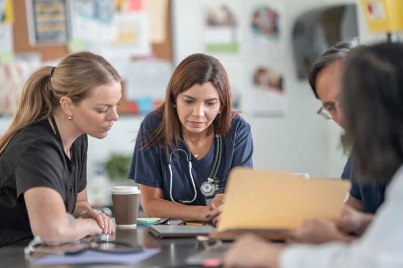 A diverse group of healthcare professionals, including a nurse and doctors, gathers around a table to discuss patient care. They review documents and share ideas in a bright clinic setting.