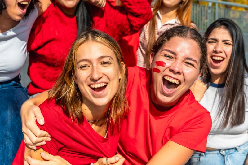 Young adult friends in red shirts cheering passionately at a sunny stadium, face paint and big smiles conveying joy, camaraderie and vibrant celebration of their team
