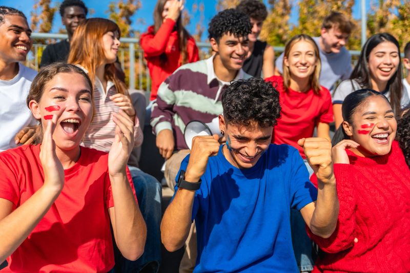 Excited multi-ethnic teens and young adults with painted faces cheering and celebrating a sports victory together on stadium bleachers, smiling, waving, and showing team spirit