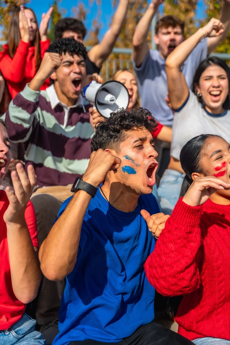 Diverse group of young adult fans, some wearing face paint and holding a megaphone, actively cheering and expressing their excitement during a sport event in a stadium