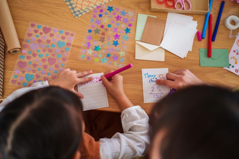 Overhead view of a mother and her young daughter creating handmade Christmas cards at a cozy wooden table. Surrounded by colorful paper, stickers, ribbons, and envelopes, they write cheerful holiday messages together, capturing the warmth and creativity of the festive season.