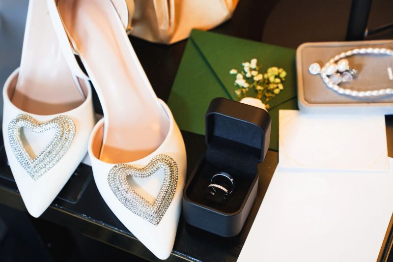 Beautiful white high-heeled shoes with heart designs sit beside a wedding ring in a black box. A green envelope and a pearl necklace complete the charming bridal setup.