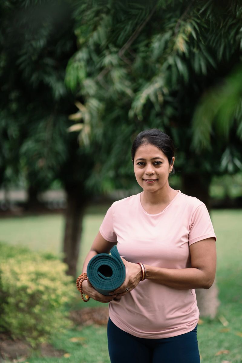 Portrait of a smiling woman standing in a lush green park while holding a rolled yoga mat. Dressed casually in a light pink t-shirt, she reflects calmness, balance, and motivation for a healthy and mindful lifestyle.