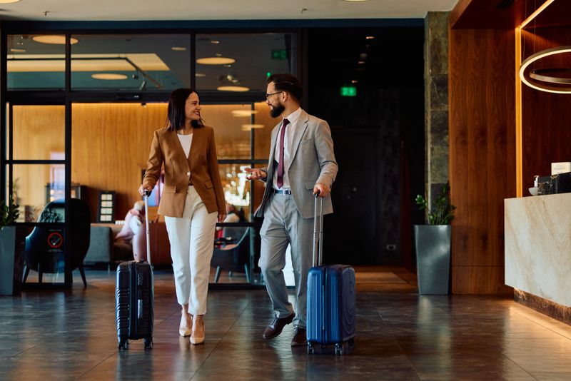 Two business professionals walking through a modern hotel lobby, carrying luggage, and engaging in conversation about their corporate travel while checking in