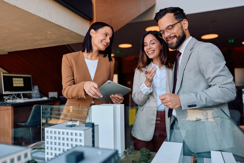 Three happy business professionals wearing formal wear are discussing an urban development project, looking at a scale model and using a digital tablet in a modern office