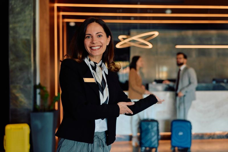 Smiling hotel receptionist standing in a modern lobby, extending her arm in a welcoming gesture, with guests and suitcases visible at the check-in desk in the blurred background