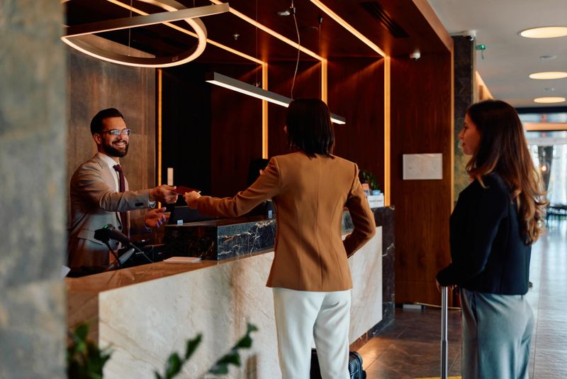 Hotel front desk agent smiles while handing a room key card to a guest checking in, with another guest waiting nearby in an elegant modern lobby during arrival