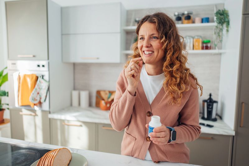 A smiling woman is taking her vitamins in a contemporary kitchen, emphasizing personal well-being and health amidst a backdrop of fresh produce and modern decor.