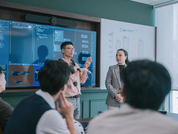 A man presenting data on screens to an attentive group in a meeting room.