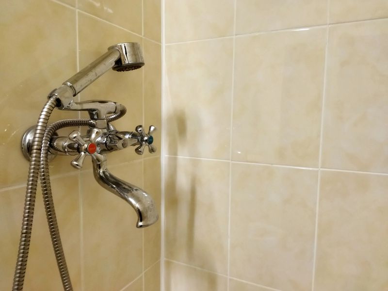 A modern shower faucet is showcased in a bright bathroom environment. The faucet features a stylish metal finish with distinct knobs for hot and cold water. The flexible showerhead with a sleek design adds a contemporary touch. The tiles surrounding the faucet are light-colored, creating a clean and inviting atmosphere. This setup highlights both functionality and aesthetic appeal in bathroom design.