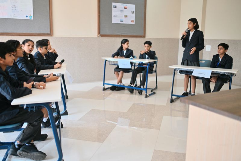 Student teams, dressed in formal school blazers, face each other for a quiz. One student stands with a microphone to answer on behalf of her team, while the other teams watch closely, ready with their own mics and buzzers to respond in the next turn.