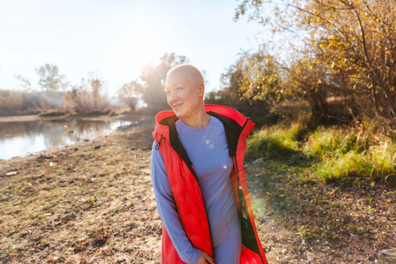 A woman with a shaved head stands by a sunlit lake in autumn, wearing a bright red vest. She smiles peacefully, capturing a moment of outdoor fitness, nature, and calm resilience.
