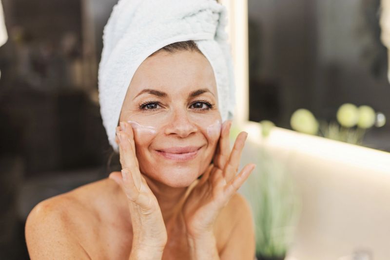 A cheerful woman with a white towel wrapped around her head applies facial cream, smiling at the mirror. The scene conveys self-care, beauty, and a relaxing skincare routine in a modern bathroom.