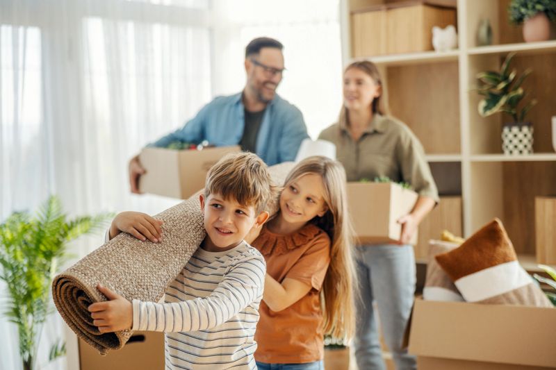 Happy family of four moving to a new house. Parents and children carrying packing boxes and a rolled rug