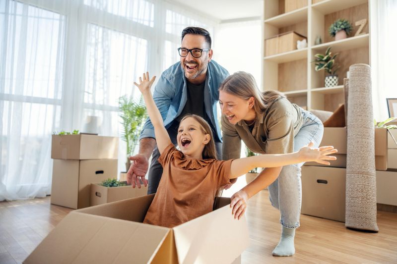 Parents pushing their happy daughter in a cardboard box, celebrating a fun moment while moving into a new house