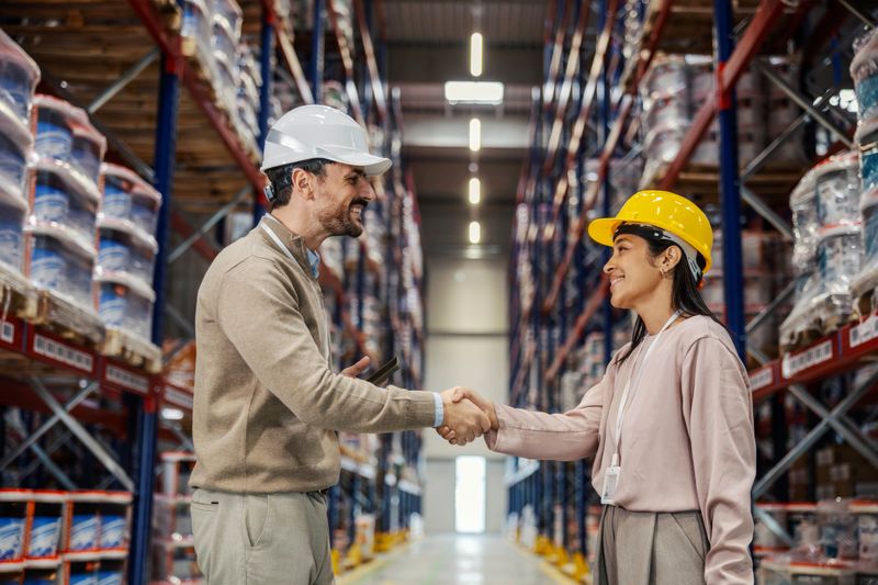 Logistics partners shaking hands in a large warehouse after reaching a business agreement. They are wearing hard hats