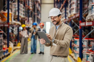 Warehouse worker wearing a hard hat using a tablet with colleagues in the background.