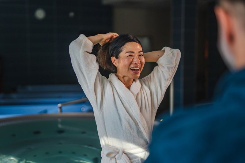 Woman in a bathrobe laughing joyfully while pulling her hair back, sharing a happy and relaxing moment with a partner at a luxury spa or health resort