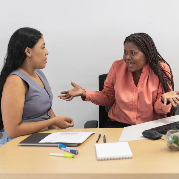 Two women engaged in an expressive conversation at a desk with office supplies.